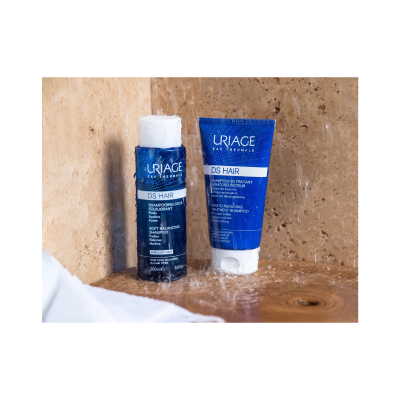 Dois frascos de champô azul da marca Uriage DS Hair numa bancada de madeira e azulejo bege com água a cair