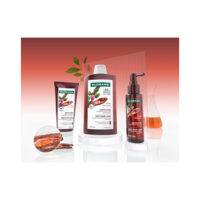 Produtos Klorane anticqueda em frascos castanhos com rótulos brancos e verdes sobre superfície branca e fundo degradê laranja.