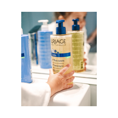 Garrafa de óleo de limpeza Uriage Bebé com bomba azul na pia e mão molhada em frente a espelho