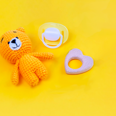 Boneco de tricô laranja em forma de urso, chupeta transparente branca e brinquedo de silicone rosa em forma de coração sobre fundo amarelo.