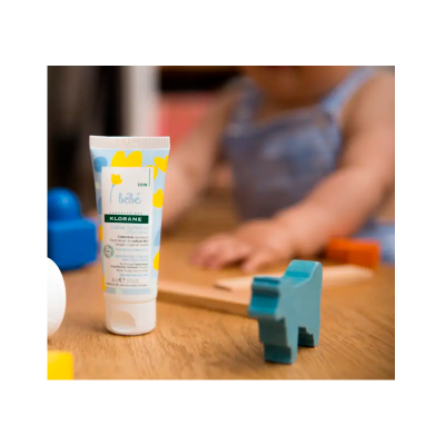 Creme para bebé Klorane com design colorido numa mesa com brinquedos e criança ao fundo