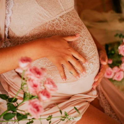 Mulher grávida com vestido rosa e renda branca, rodeada de flores cor-de-rosa