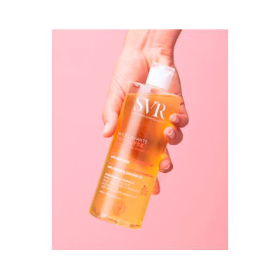 Frasco de óleo de limpeza SVR Topialyse amarelo segurado por mão sobre fundo rosa