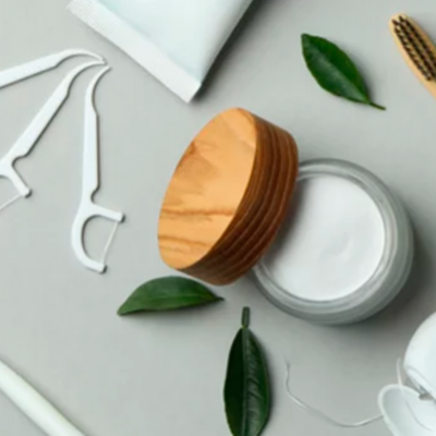 Produtos para cuidados orais com pote branco e tampa de madeira, palitos dentais, recipiente branco e folhas verdes sobre fundo cinza.