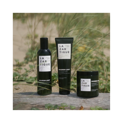 Três produtos de cuidado capilar Lazar Tigue em embalagens pretas com rótulos brancos, dispostos sobre madeira com vegetação atrás.