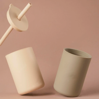 Três recipientes cilíndricos em tons bege e taupe com acabamento fosco sobre fundo rosa claro