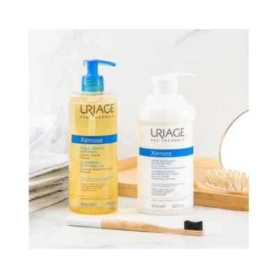 Produtos URIAGE Xémose com óleo de limpeza amarelo e creme branco, escova de dentes de madeira e toalhas cinza
