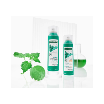 Dois frascos verdes de champô seco KLORANE com urtiga, folha de urtiga fresca e frasco de vidro com líquido verde.