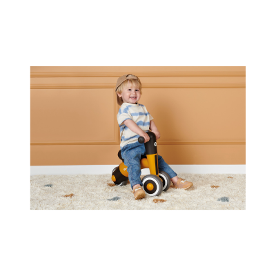 Criança numa bicicleta de balanço amarela e preta num tapete branco com fundo laranja