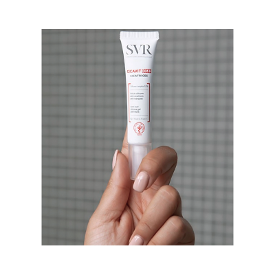 Tubo branco de gel anti-cicatrizes SVR Cicavit DM+ segurado por mão com unhas nude num fundo de azulejos cinzentos.