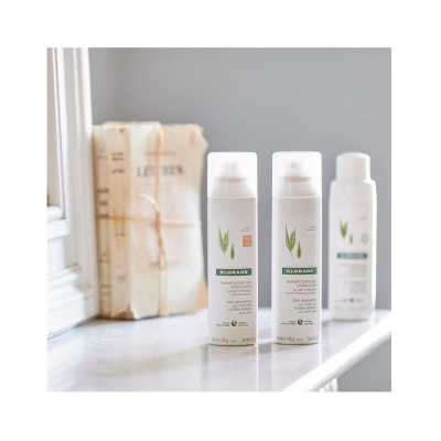 Dois frascos de champô seco Klorane em superfície branca com livros ao fundo