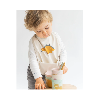 Criança com babete bege e tigelas coloridas numa mesa de madeira