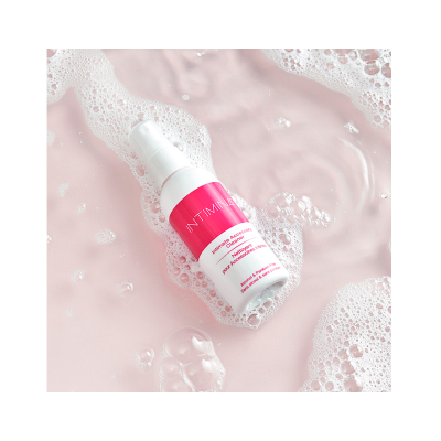 Frasco spray branco com rótulo rosa Intimina sobre fundo rosa com espuma e água