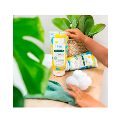 Gel de banho Klorane bebé com extrato de camomila numa embalagem amarela, azul e branca em balcão de madeira, com toalha, brinquedo e planta