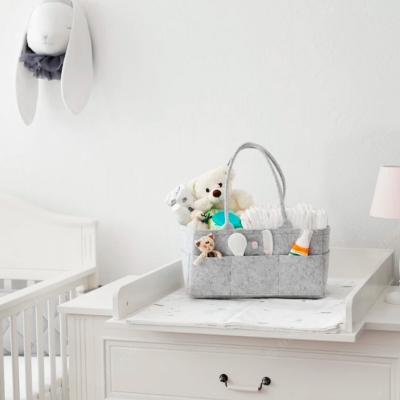 Cesto organizador cinzento com fraldas e brinquedos numa cómoda branca para muda fraldas com lâmpada rosa e decoração de coelho na parede.