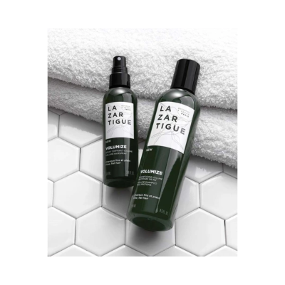 Frascos verdes escuros de produtos LAZARTIGUE Volumize sobre azulejos brancos com toalha branca ao fundo