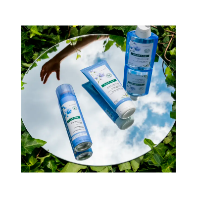 Produtos de cosmética azul e branca sobre espelho refletindo céu e mão, com folhas verdes ao redor