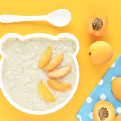 Prato em forma de urso com mingau e fatias de pêssego, colher branca, pêssegos inteiros e cortados, toalha azul com bolinhas brancas numa superfície amarela