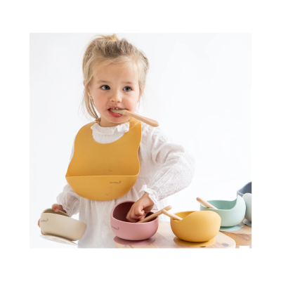 Criança com babete de silicone amarelo e tigelas coloridas com colheres de madeira.