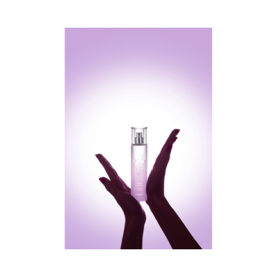 Frasco translúcido de spray rosa segurado por duas mãos femininas com fundo lilás