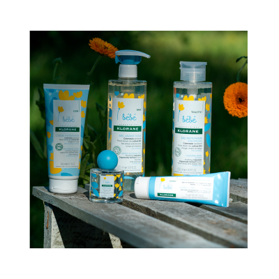 Produtos de cuidados para bebé Klorane em embalagens brancas, amarelas e azuis, dispostos numa superfície de madeira ao ar livre com flores alaranjadas ao fundo.