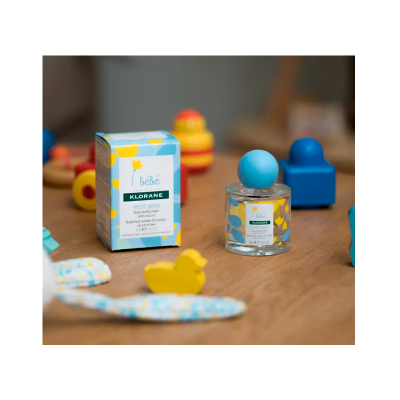 Perfume infantil KLORANE BEBE PETIT BRIN com frasco e caixa em mesa de madeira rodeado de brinquedos coloridos