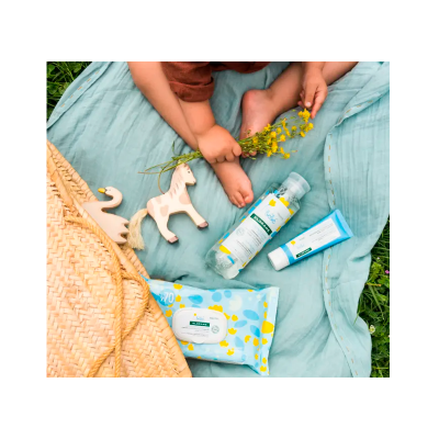 Produtos para bebé sobre tecido azul claro com brinquedos de madeira e saco de palha, crianças segurando flores amarelas