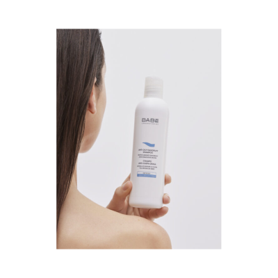 Shampoo anti caspa oleosa BABE Laboratorios em embalagem branca segura por mulher de costas com cabelo castanho