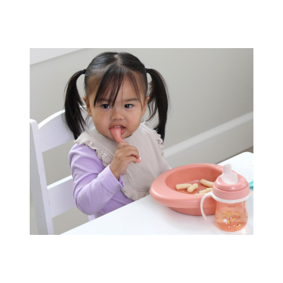 Criança com babete a mastigar colher junto a prato e copo para crianças.