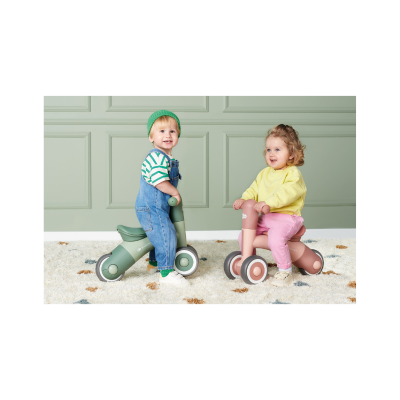 Duas crianças pequenas em bicicletas de corrida sem pedais plástico verde e rosa num tapete branco e parede verde