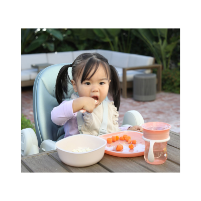 Criança a comer cenouras em cadeira alta com prato e copo cor-de-rosa numa mesa ao ar livre