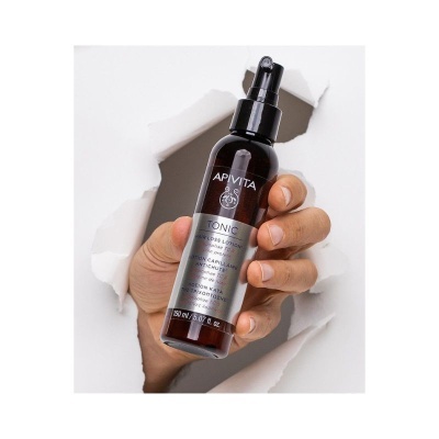 Frasco de loção capilar APIVITA TONIC com pulverizador preto segurado por uma mão a sair de papel rasgado branco