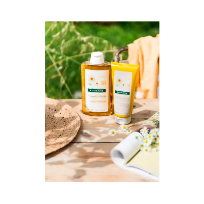 Produtos Klorane em frasco e tubo amarelo sobre mesa de madeira com chapéu de palha e flores