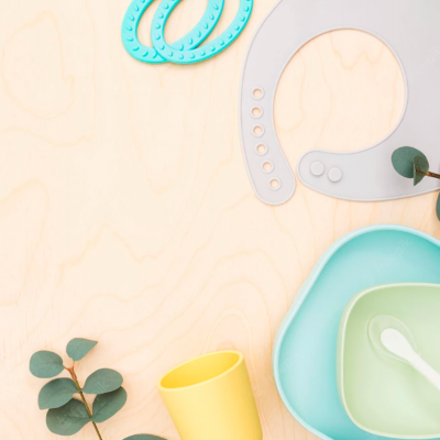 Utensílios de alimentação infantil em cores pastel com babetes, prato, copo e colher sobre superfície de madeira.