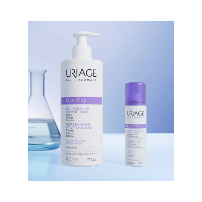 Dois frascos da linha URIAGE Gyn-Phy para higiene íntima de cor branca com detalhes roxos, um maior com doseador e outro menor em spray, em fundo azul claro com frasco de vidro