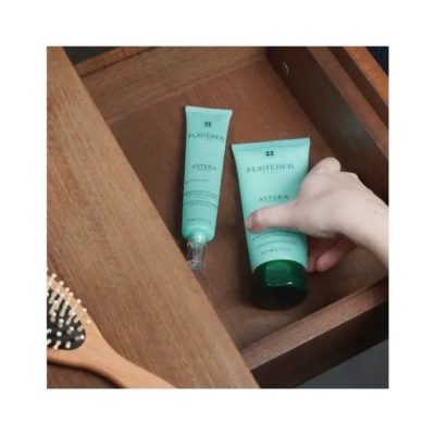 Dois tubos verdes de creme FURTERER ASTERA dentro de gaveta de madeira, mão a segurar um tubo, escova de cabelo ao lado.