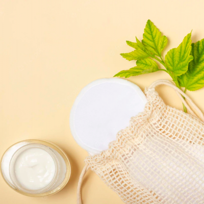 Frasco de creme aberto, discos de algodão e bolsa de malha sobre fundo bege com folhas verdes