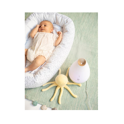 Bebé deitado em almofada branca com padrão, brinquedo polvo amarelo e difusor branco com botão de madeira num tapete verde