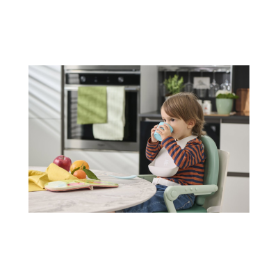 Criança sentada numa cadeira alta a beber de um copo azul numa cozinha com frutas e livro sobre a mesa.