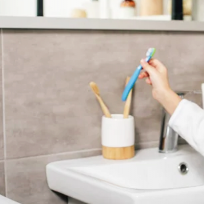 Mão segurando escova de dentes azul ao lado de sanita branca e copo com escovas de dentes numa casa de banho.