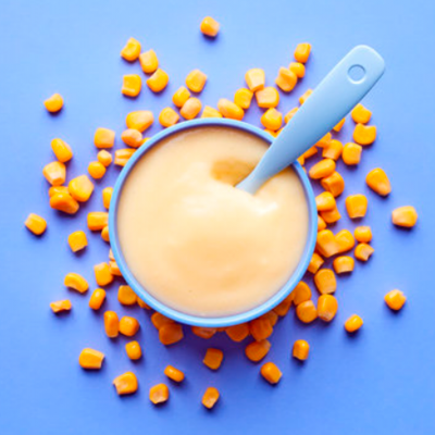 Taça azul com papa amarela e colher azul rodeada por grãos de milho amarelos