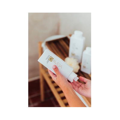 Frasco branco de produto segurado por mãos em banco de madeira com outros frascos e flores ao fundo