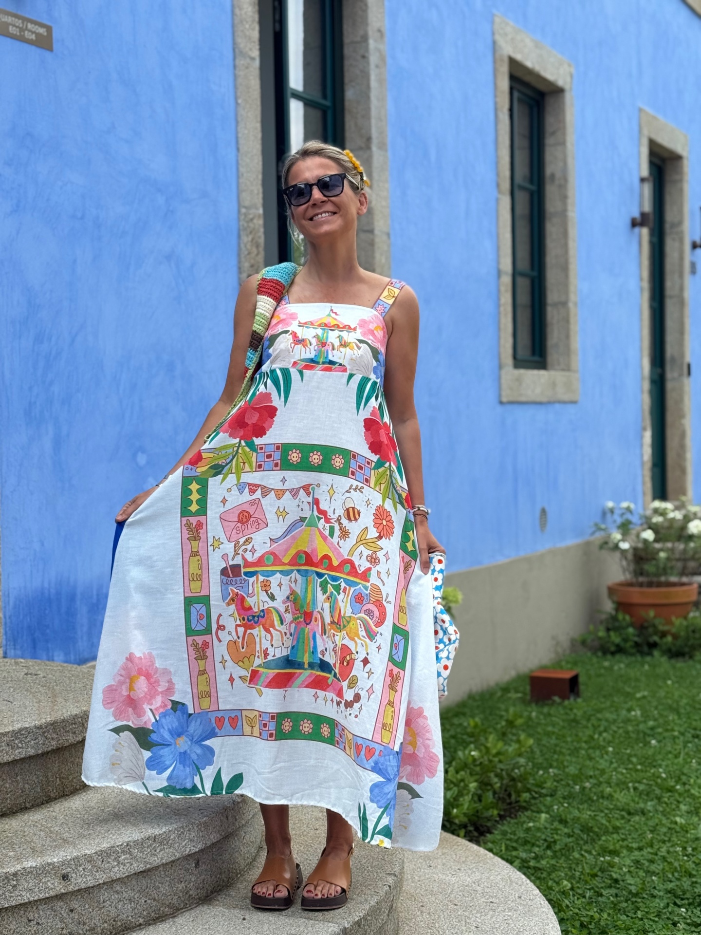 Mulher com vestido longo estampado colorido ao ar livre junto a parede azul e janelas.
