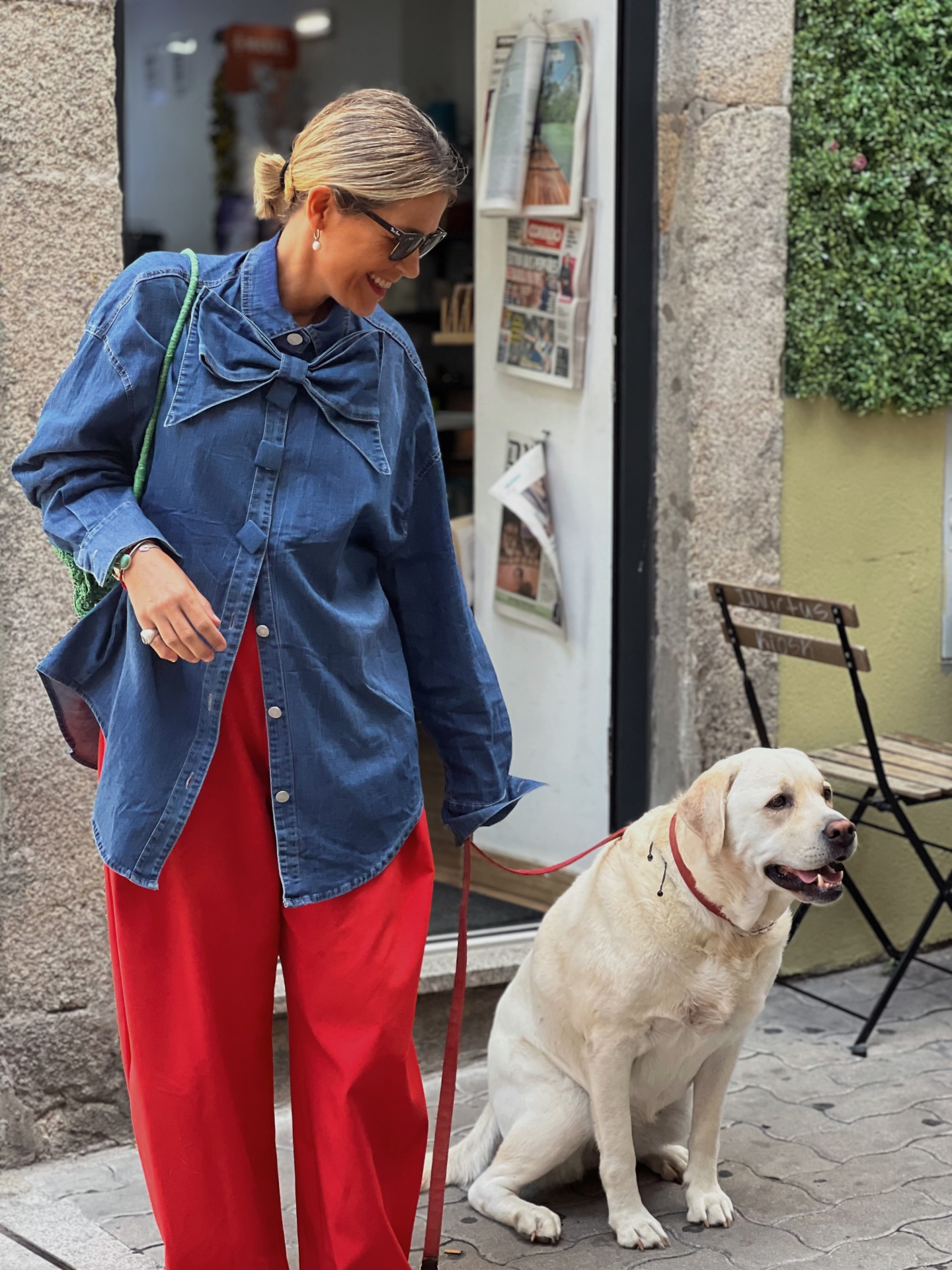 Mulher com camisa azul de denim com laço, calças vermelhas, óculos escuros e cão labrador creme sentado