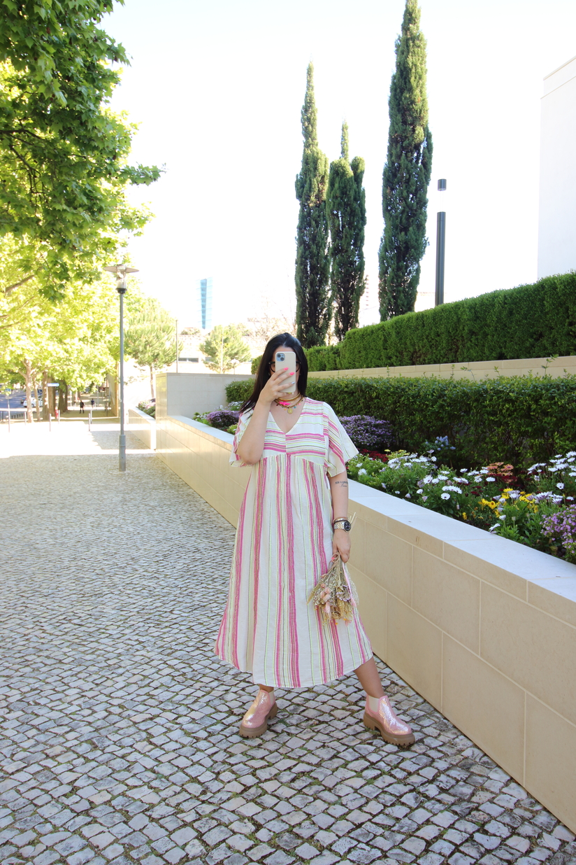 Mulher com vestido de algodão às riscas verticais rosa e branco, sapatos rosa, segurando flores secas numa rua com calçada portuguesa e vegetação
