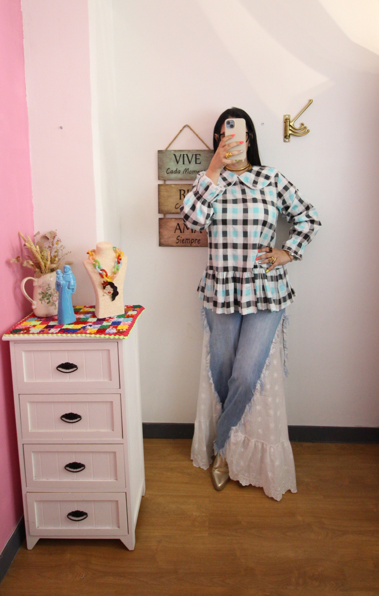 Pessoa com blusa xadrez, calças de ganga e sapatos dourados, tirando selfie junto a cómoda e placa na parede.