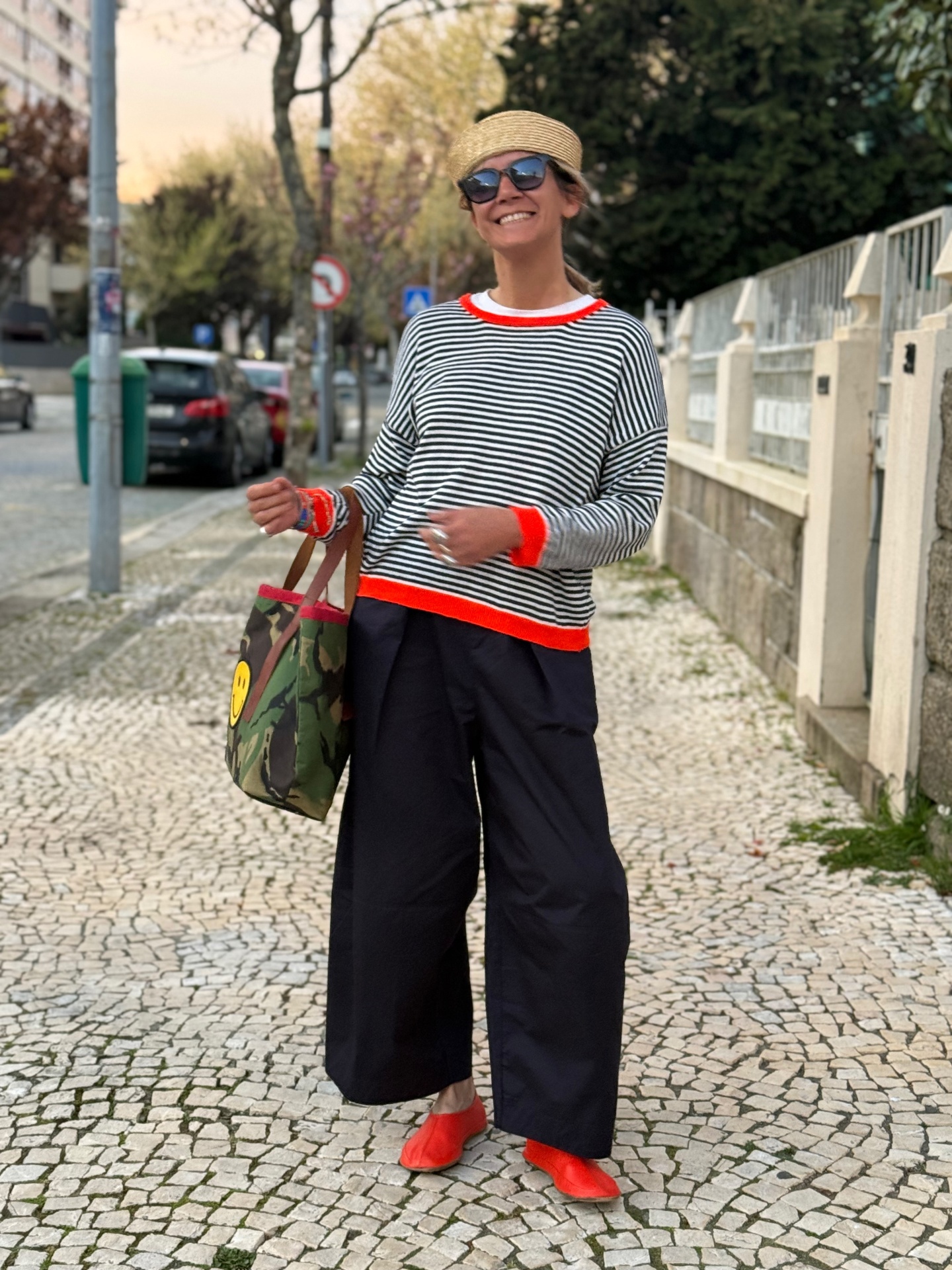 Mulher com camisola riscada, calças largas, sapatos vermelhos e bolsa camuflada numa calçada.
