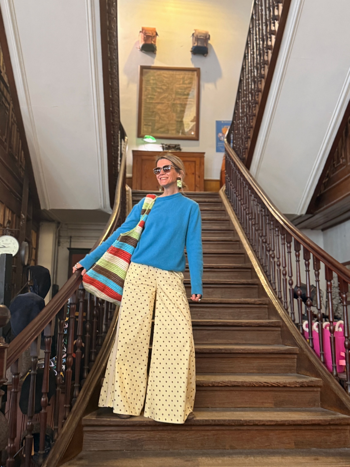 Mulher com camisola azul, calças largas bege com bolinhas e saco colorido numa escadaria em madeira