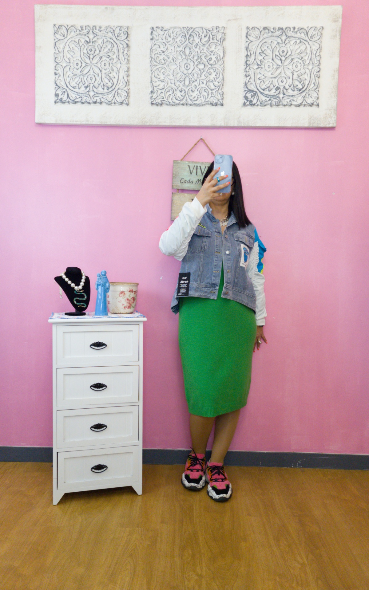 Pessoa tirando selfie com vestido verde e colete jeans em frente a parede rosa com decoração e móvel branco com bijuterias.