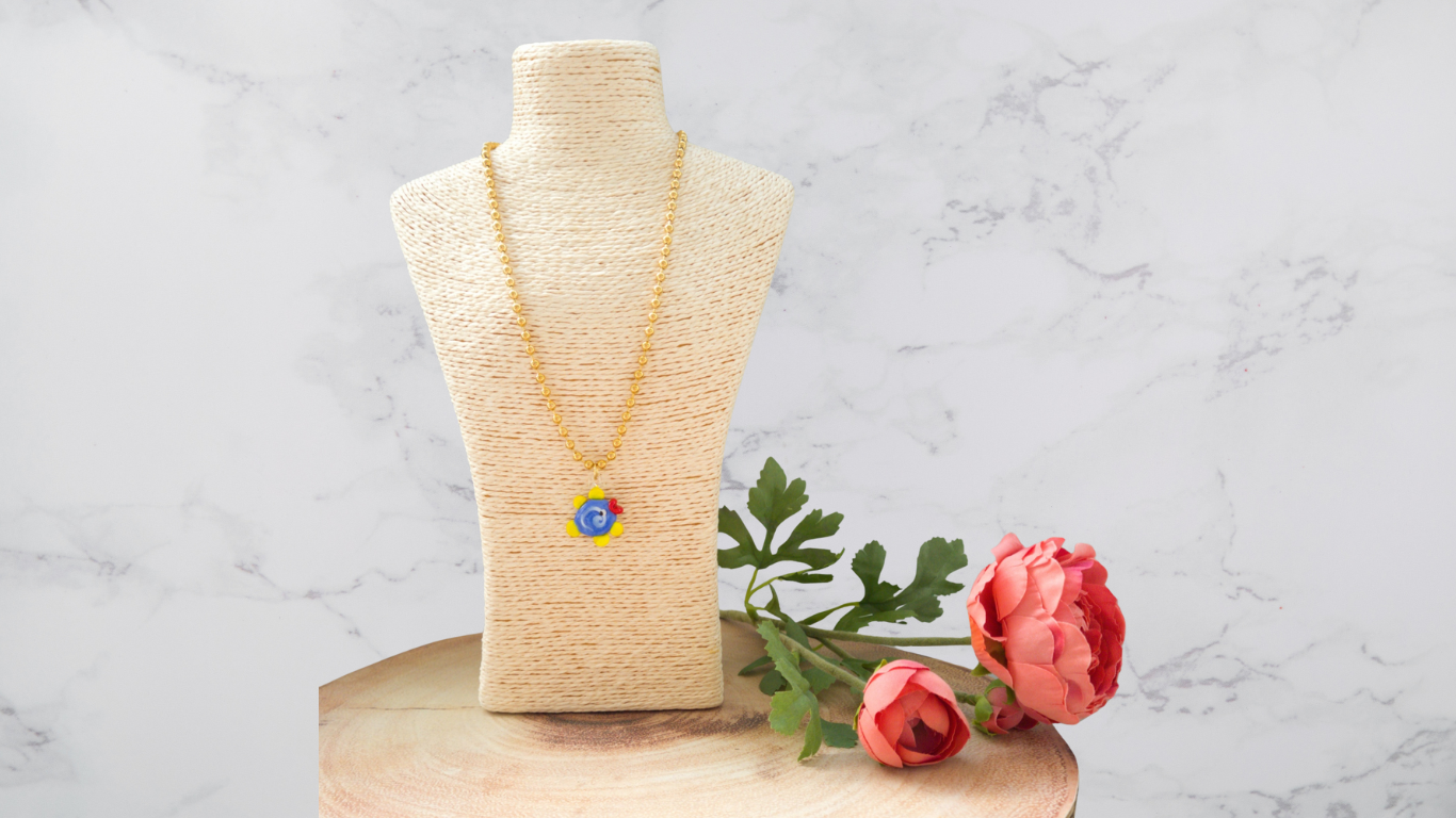 Colar dourado com pendente colorido em busto de tecido e flores rosas ao lado